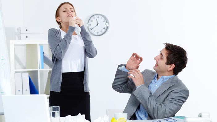 Woman sneezing at work while coworker looks at her.