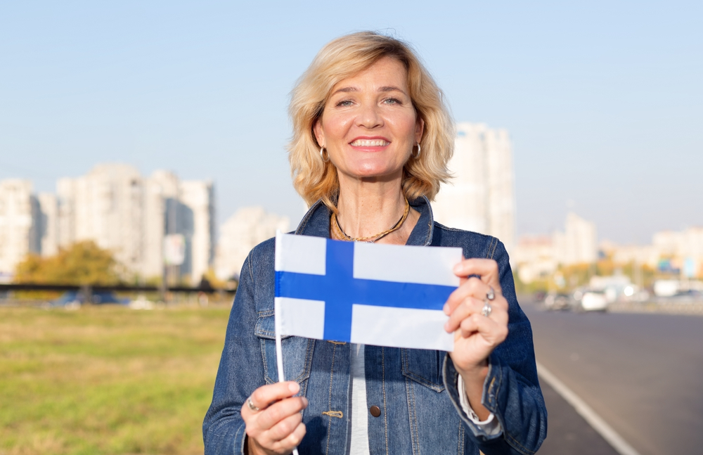 Happy mature woman with flag of Finland standing against the background of a city street and blue sky.