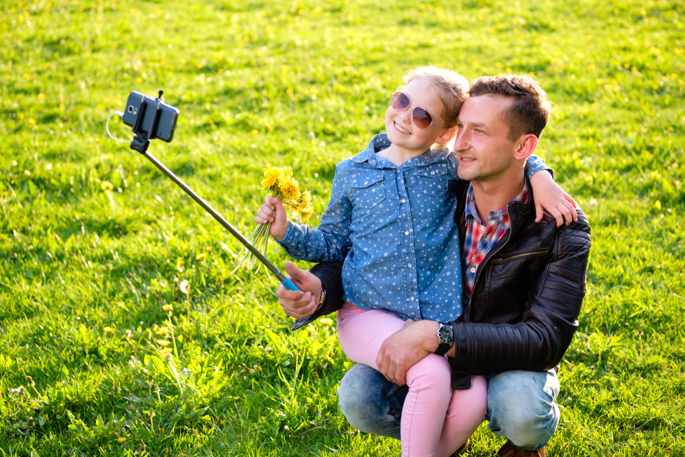 Happy Dad and little daughter doing picture by smartphone use selfie stick in summer park.
