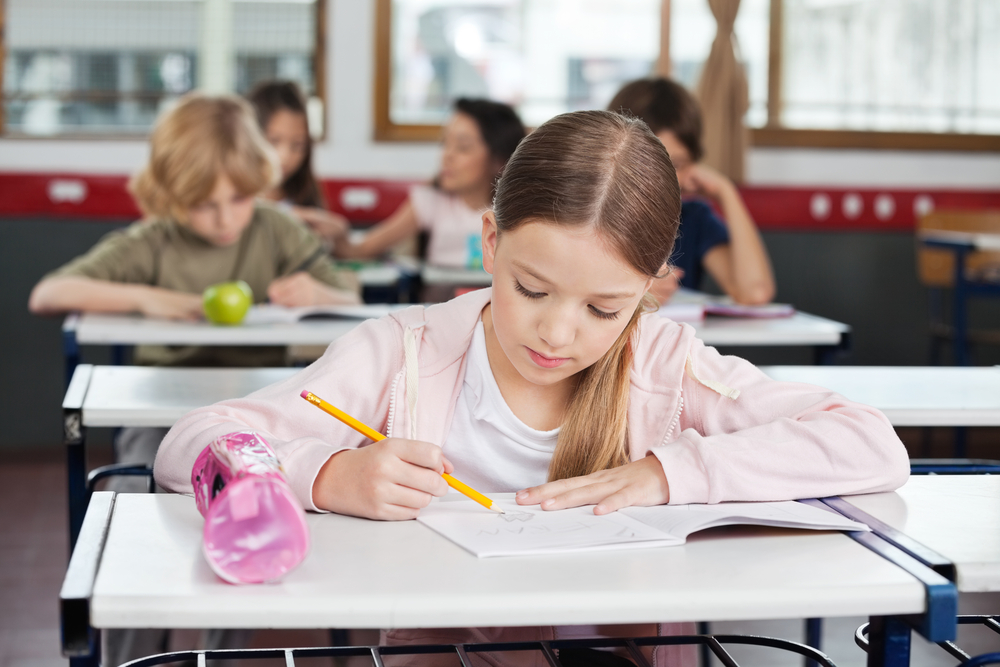 Cute little schoolgirl drawing in book with classmates in background at classroom