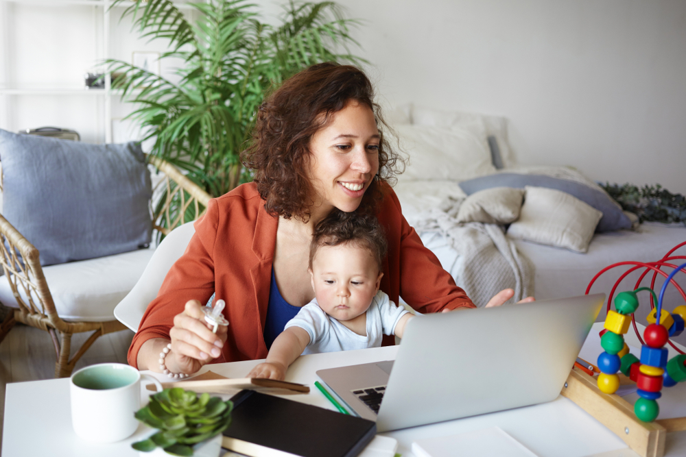 Attractive young dark skinned woman working at desk at home using laptop, holding baby on her lap. Portrait of smiling mother writing post on moms blog while her infant son playing with toy s